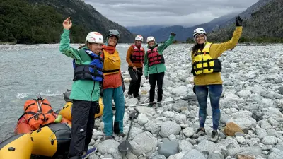 Five professional female mountaineers on a riverbank in Patagonia wave and laugh at the camera.