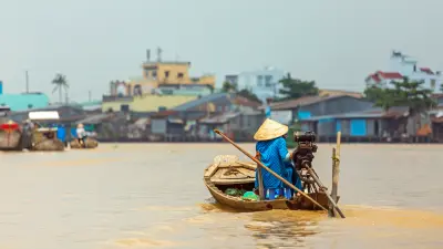 A person in a traditional conical hat navigates a wooden boat on a river. A settlement is visible in the background.